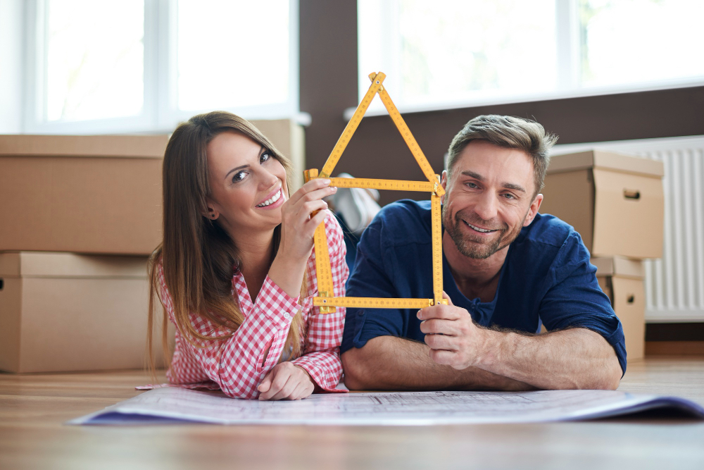 772025 - Vivir e Invertir en Canarias - Haznoticia.com A couple smile while lying on the floor with a blueprint and a yellow triangular ruler forming a house shape, surrounded by moving boxes.
