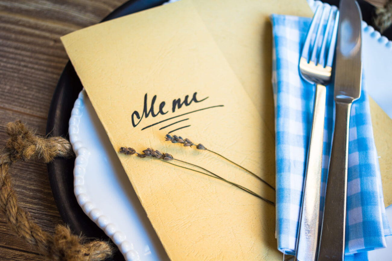 Table setting in rustic style with Menu - Haznoticia.com Yellow menu on a white plate with a blue checkered napkin and silverware visible on a rustic table with lavender sprigs nearby.
