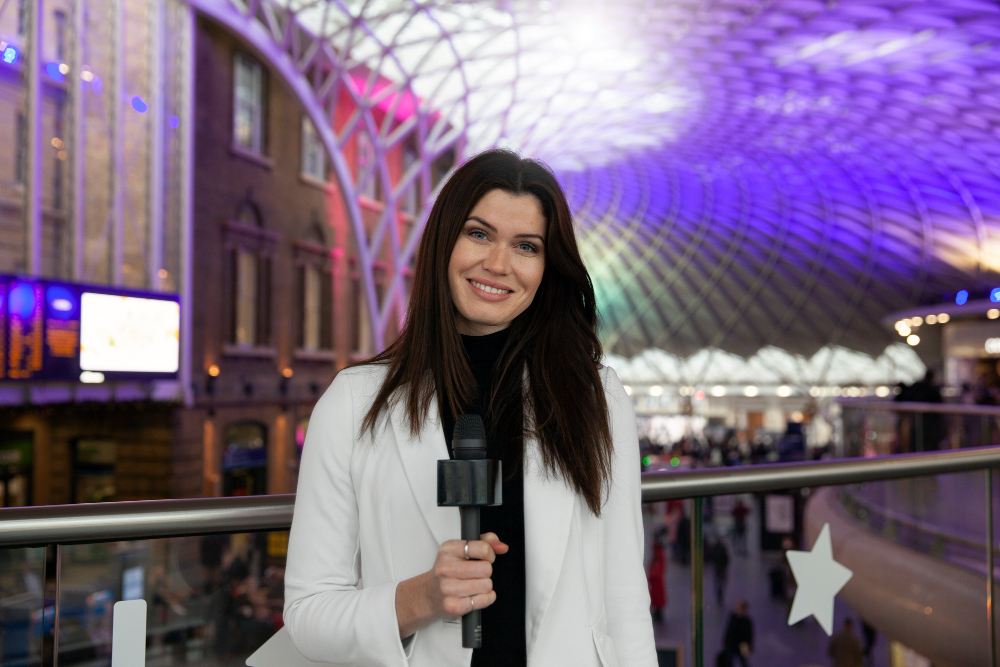 772702 - Marina Durán - Haznoticia.com Female reporter in a white blazer holding a microphone, standing in a colorful, modern indoor station.