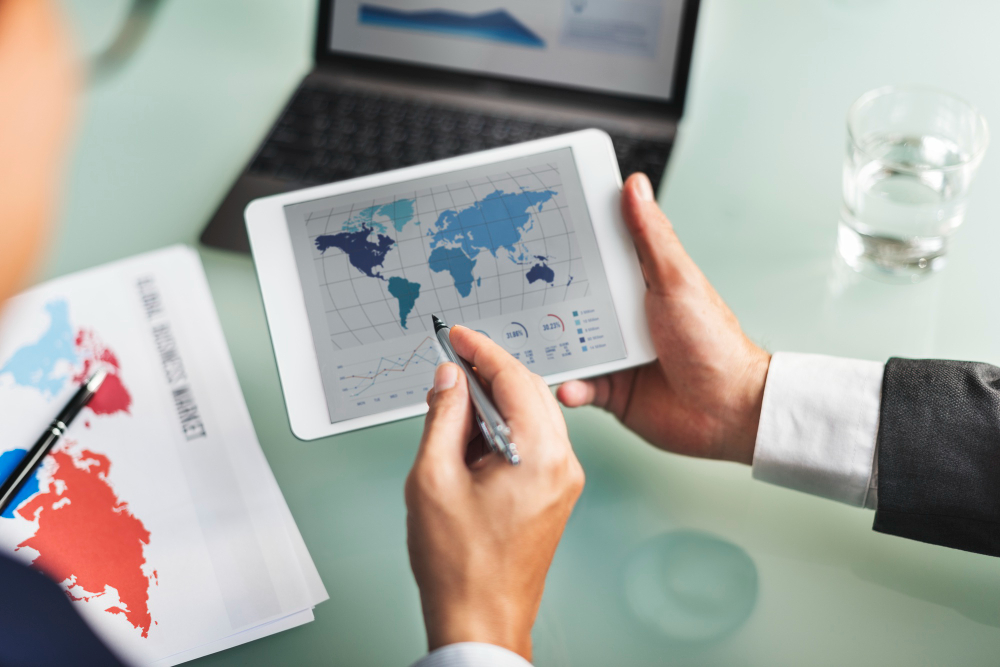772927 - Ledesma International - Haznoticia.com Person holding a tablet displaying a world map and charts while writing with a pen at a glass-topped desk.
