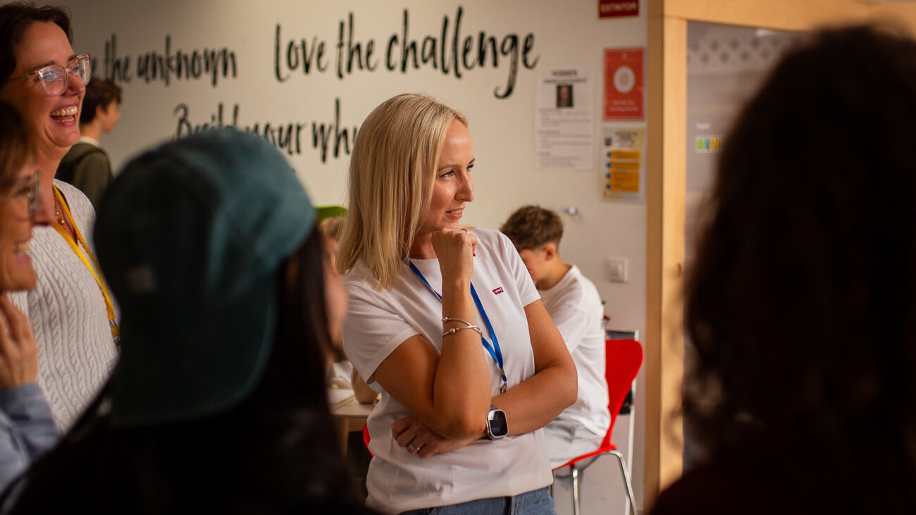 learnlife - IP - Haznoticia.com Smiling woman with blonde hair in a white shirt and blue lanyard listens thoughtfully at a group event, wall text says 'love the challenge'.