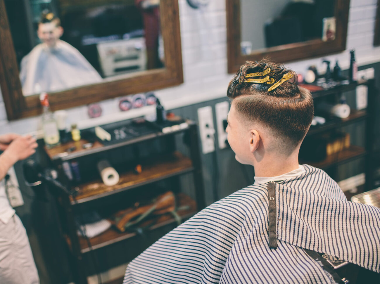hairstylist and plesed man client with haircut looking in mirror - Haznoticia.com Barber cutting a customer's hair in a vintage-style shop, with striped cape and yellow hair clips securing the top.