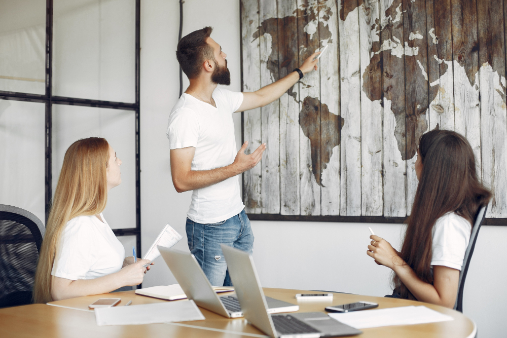 775068 - Manuel Vida - Haznoticia.com Man stands and points at a world map mural on the wall while two women listen in a conference room.