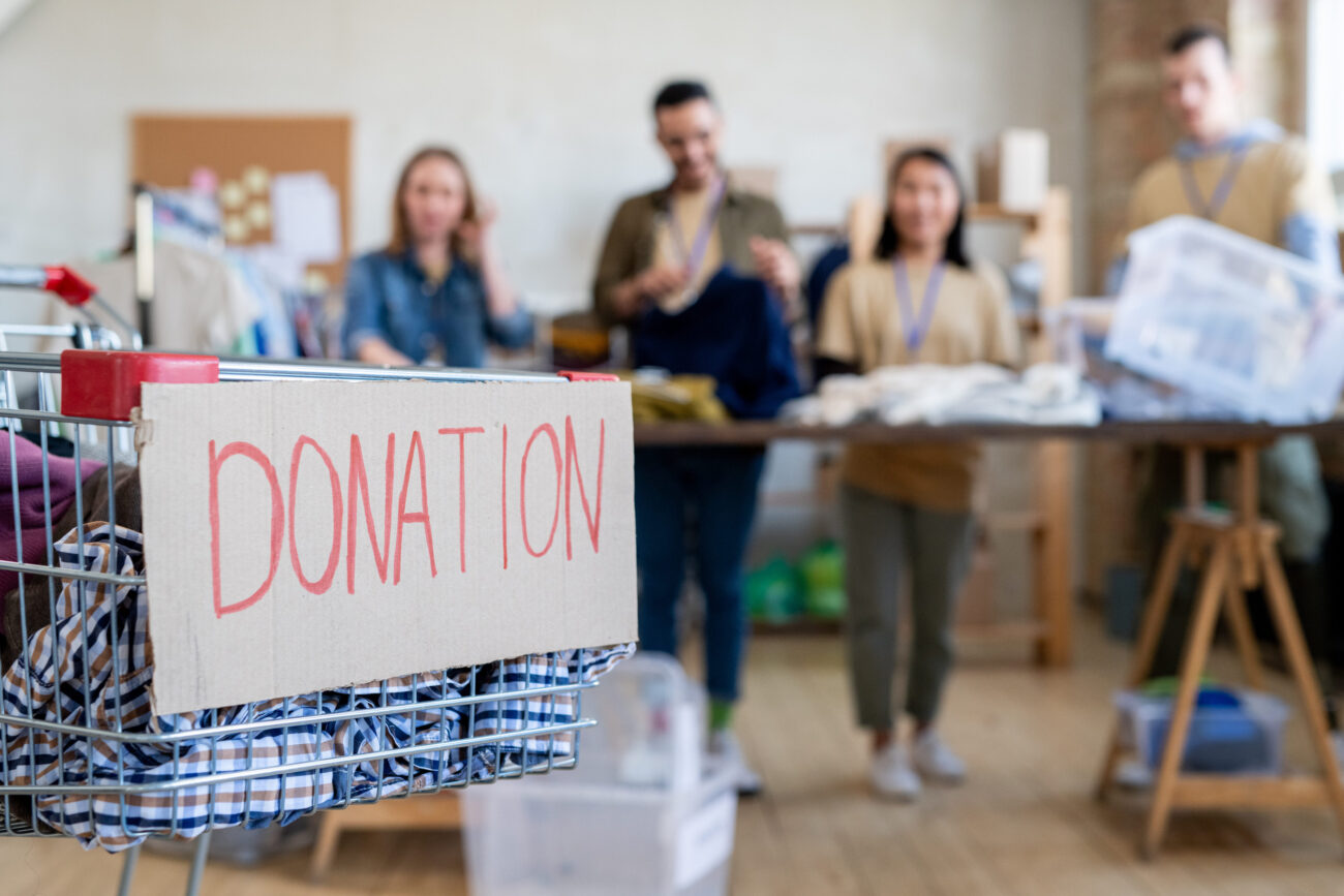 Shopping cart with donation clothes against row of volunteers standing by table - Haznoticia.com Cardboard sign reading DONATION on a clothing donation cart with folded clothes, group of people blurred in a donation drive background.