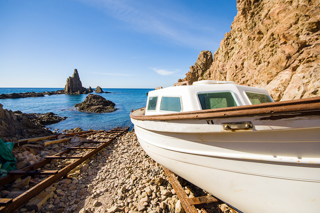 Cabo de Gata in Almeria Spain - Haznoticia.com White small boat on a pebbly beach beside rusted railway tracks, with blue sea and cliff face in the background.